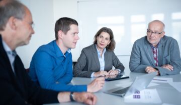 zwei Männer und eine Frau sitzen an einem Tisch und besprechen sich, dabei sind alle Augen auf den Mann links gerichtet
