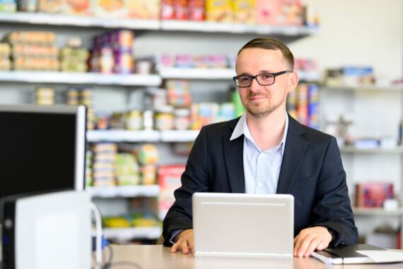 junger Handelsfachwirt mit Brille sitzt vor seinem Notebook im Einkaufsladen