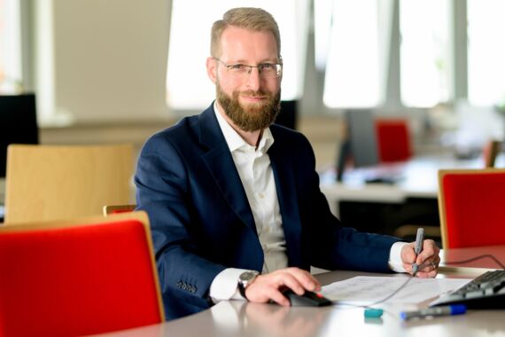 Mann sitzt am Rechner vor seinen Unterlagen und lächelt den Betrachter an
