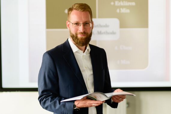 junger Mann steht im Anzug mit Unterlagen in der Hand und lächelt den Betrachter an