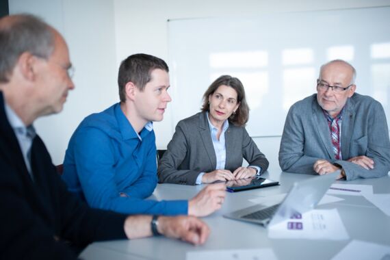 zwei Männer und eine Frau sitzen an einem Tisch und besprechen sich, dabei sind alle Augen auf den Mann links gerichtet
