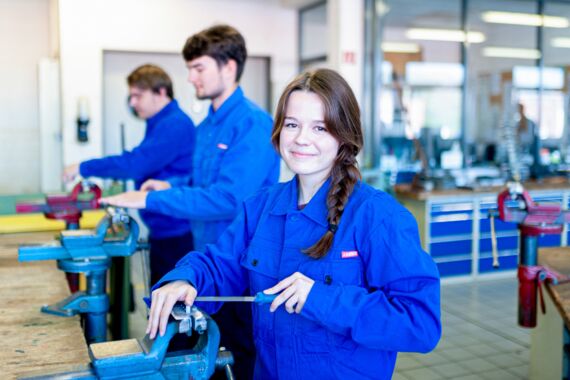 junge Frau steht in der Werkstatt am Tischschraubstock und arbeitet mit einer Werkstattfeile, dabei lächelt sie den Betrachter an