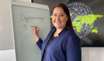 Eine Projektmitarbeiterin in einem blauen Blazer steht schreibend an einem Smartboard mit Blickrichtung Kamera.