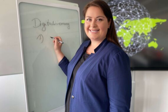 Frau steht vor einem digitalen Whiteboard und einer vernetzten Weltkarte und lächelt den Betrachter an