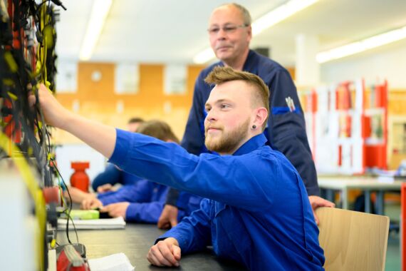junger Mann im Blaumann zeigt seine Fähigkeiten als Mechatroniker, sein Ausbilder schaut ihm dabei über die Schultern